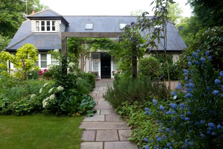 Front garden view of a stylish Surrey home with stone pathway, dense perennial borders, and climbing plants creating a sophisticated, verdant setting.