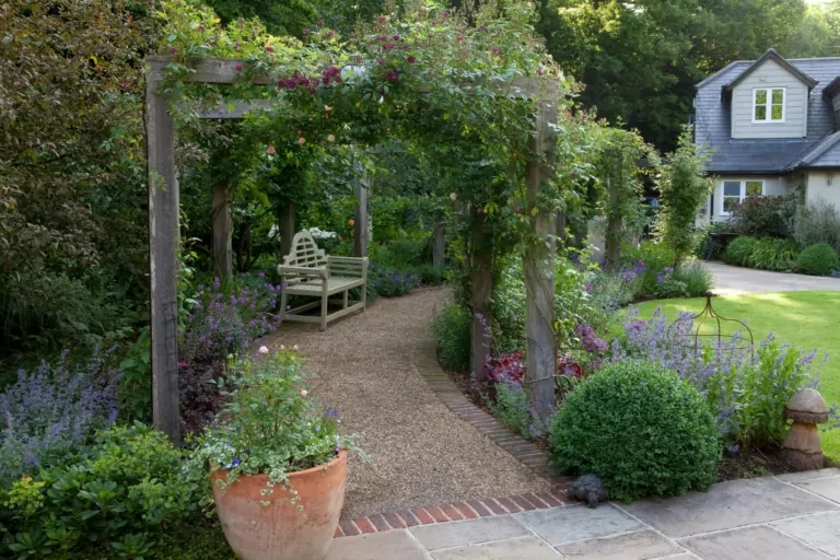 Decorative garden archway leading to a gravel pathway, flanked by terracotta pots and flowering plants, creating a welcoming entrance to a landscaped garden.