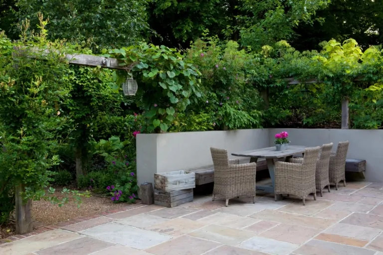 Wide-angle of paved garden terrace with table and chairs, framed by mature trees and manicured planting, offering an elegant outdoor living space.