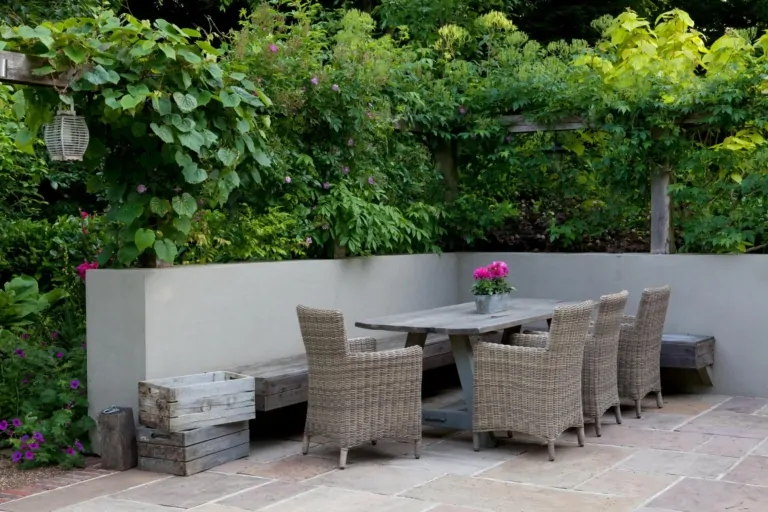 Outdoor dining terrace with wicker chairs and rustic wooden benches, set against a white garden wall and surrounded by greenery, highlighting luxury garden design.