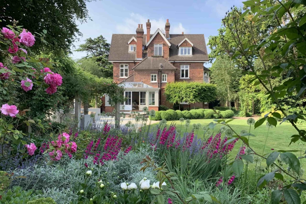Historic red-brick house with formal landscaped garden featuring colorful flower borders, manicured lawn, and blooming roses in the foreground.