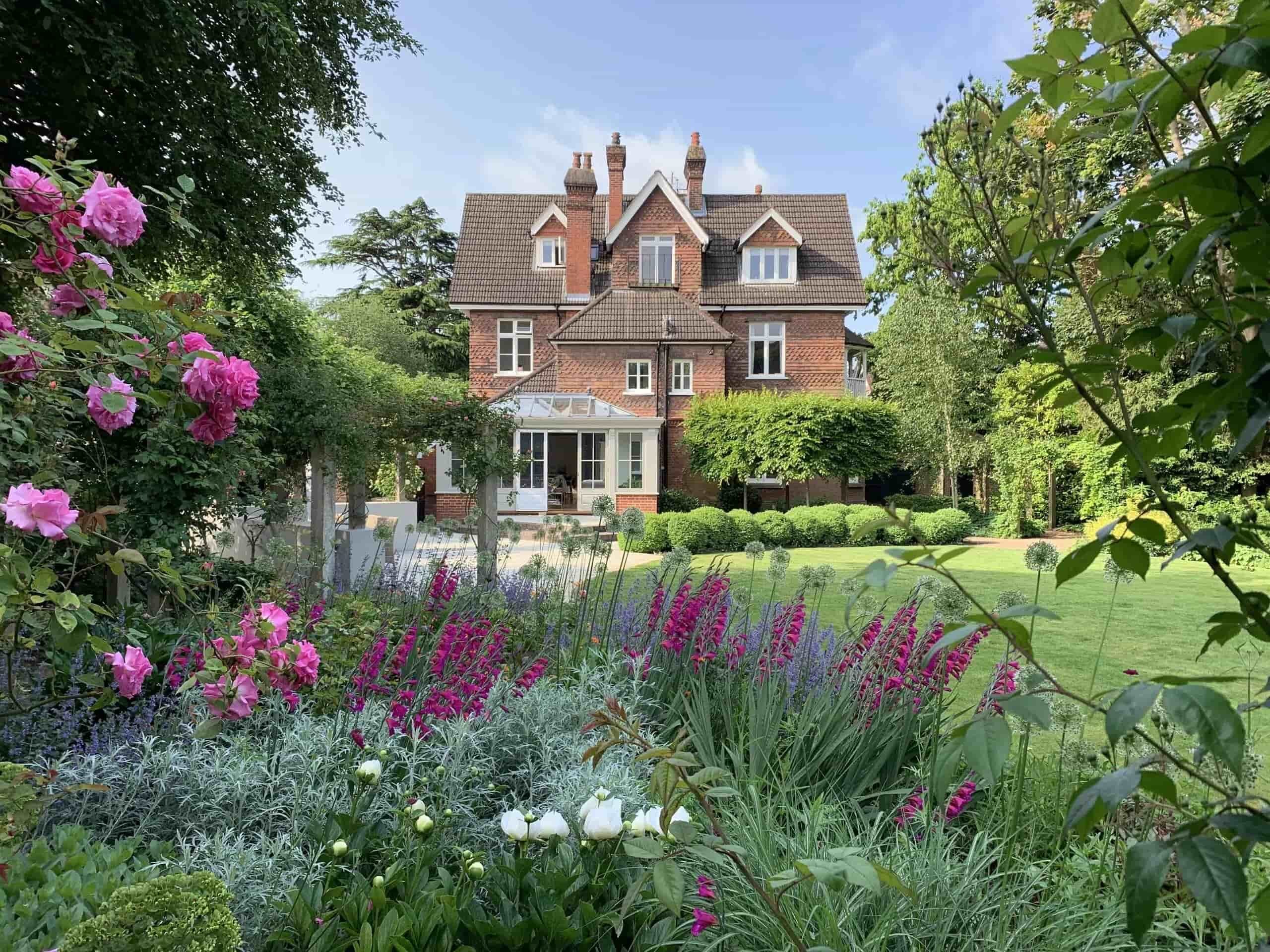 Historic red-brick house with formal landscaped garden featuring colorful flower borders, manicured lawn, and blooming roses in the foreground.