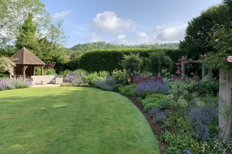 Backyard view with circular lawn, wooden gazebo, flowering borders, and manicured hedges under sunny skies.