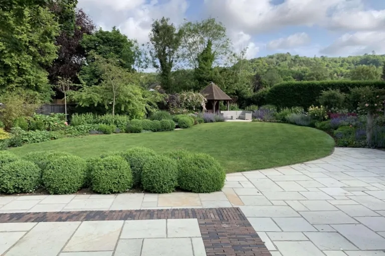 Formal garden with circular lawn, trimmed box hedges, and paved patio leading to a wooden gazebo in the background.