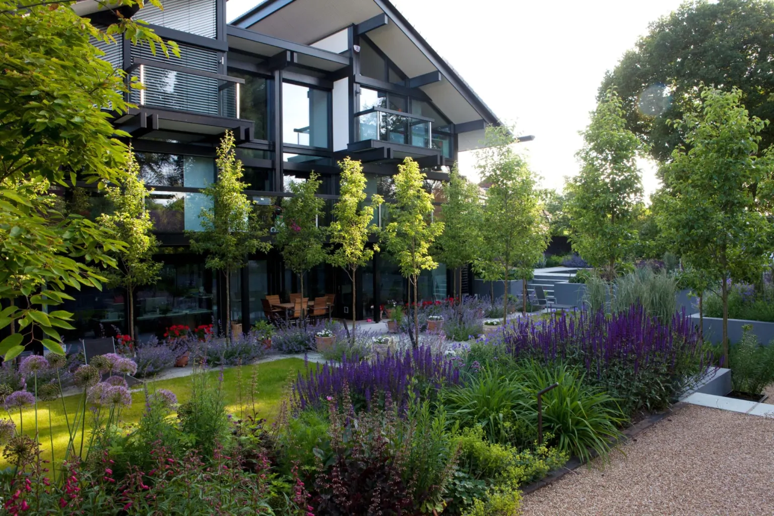 Contemporary black-and-white house with large windows overlooking a landscaped garden filled with lavender, green shrubs, and trees, creating a modern backyard oasis.