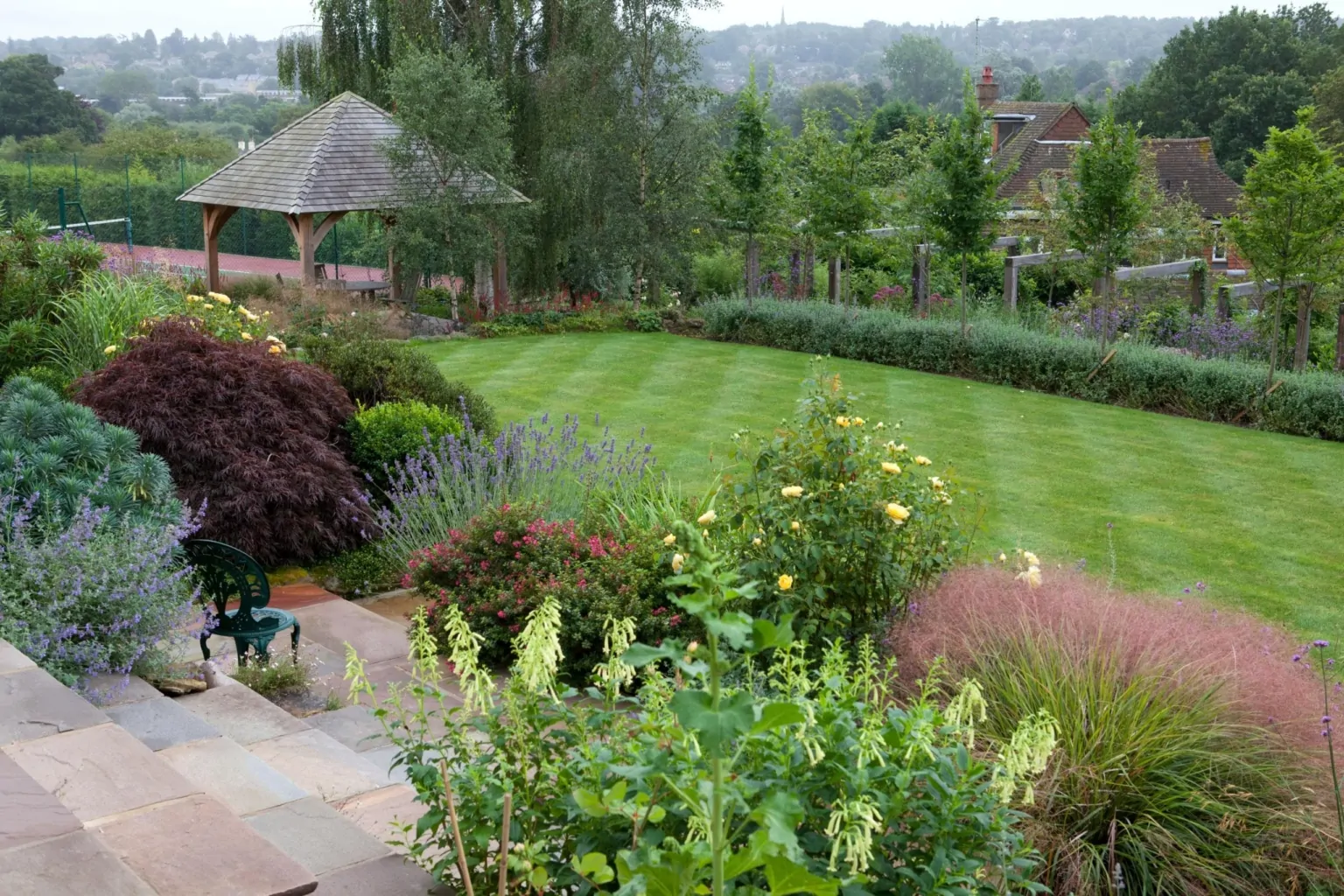 Elevated view of a landscaped garden with a manicured lawn, wooden gazebo, flowering plants, ornamental shrubs, and distant countryside views, creating a serene outdoor retreat.