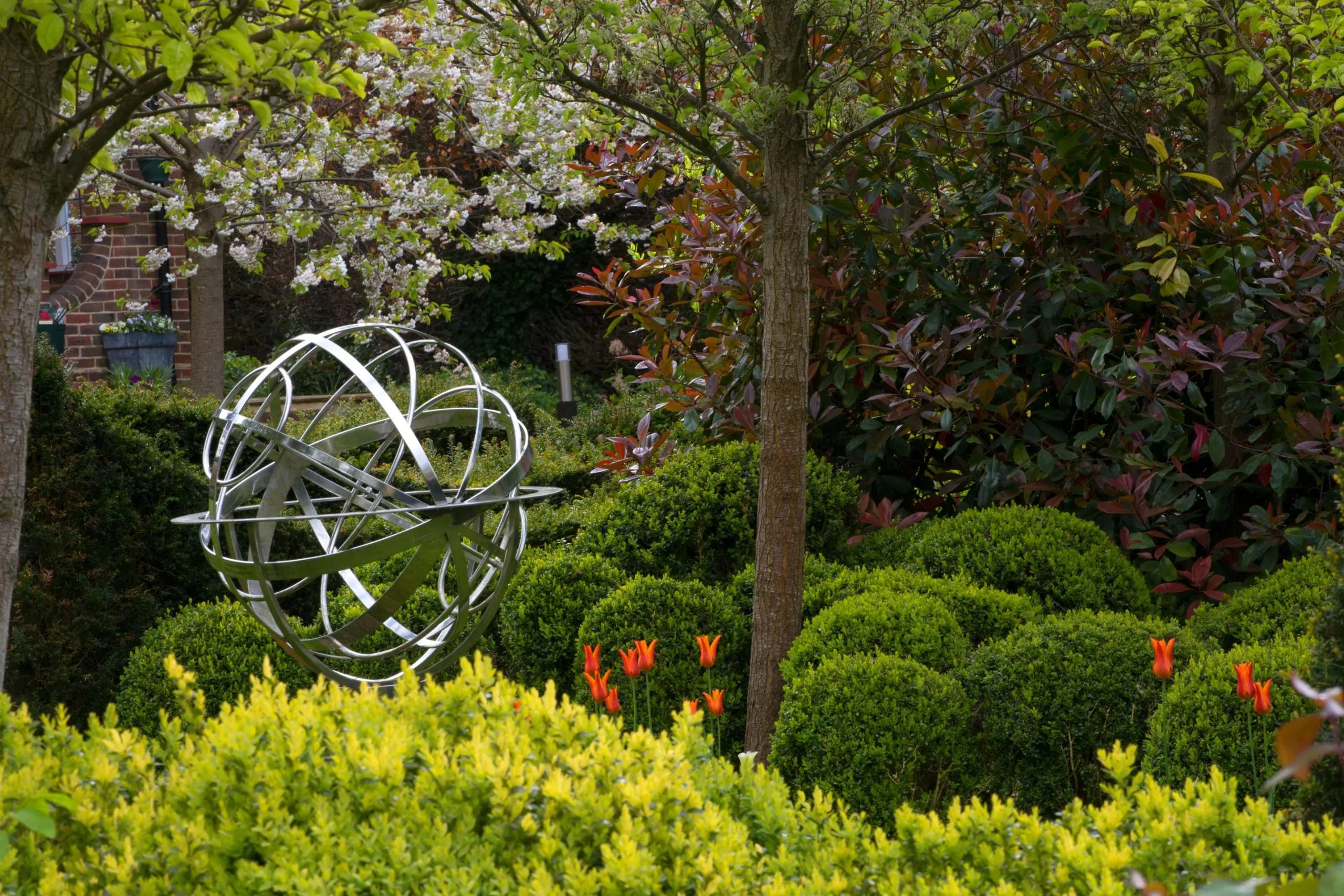 Formal garden with trimmed evergreen shrubs, orange tulips, and a decorative metal armillary sphere under trees, creating a structured and vibrant spring landscape.