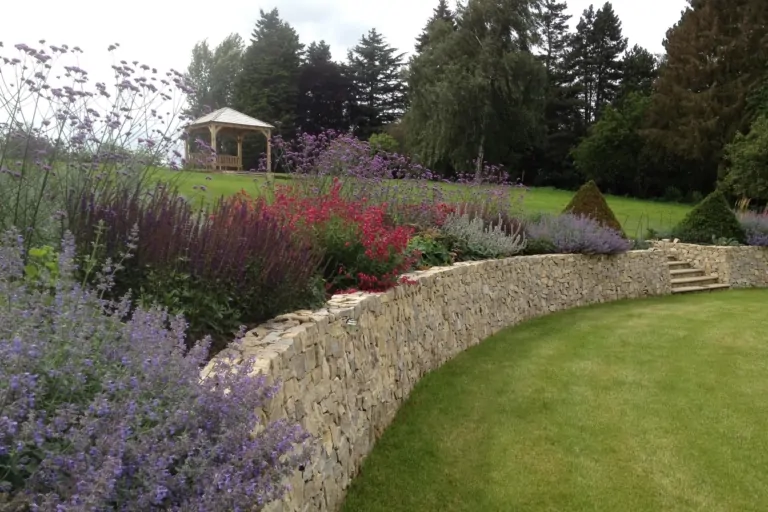 Curved stone wall with purple and red flowering plants overlooking a manicured lawn and garden gazebo.