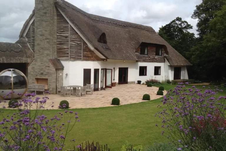 Thatched-roof house with stone patio, manicured lawn, and surrounding purple flowering garden beds.
