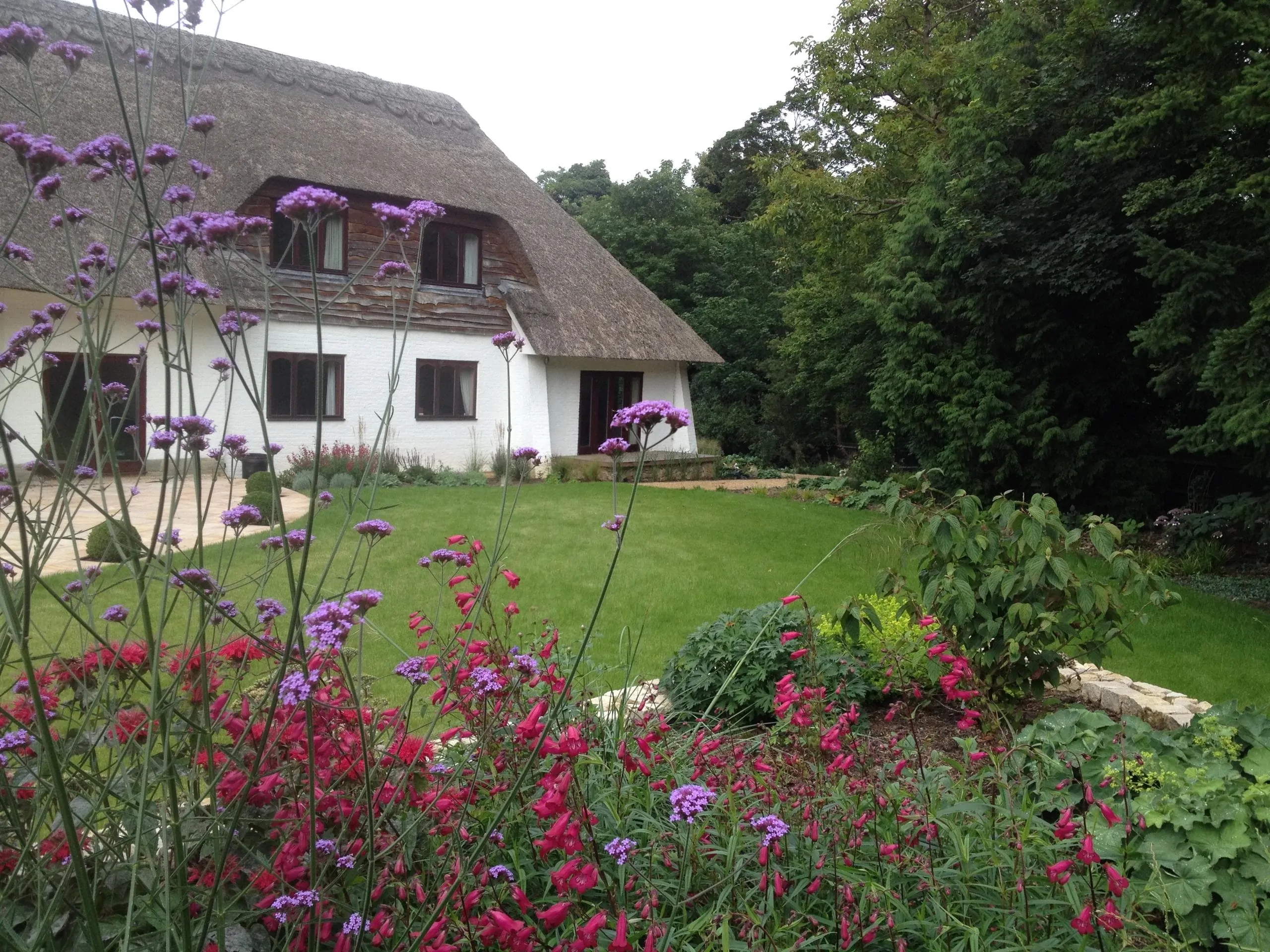 Backyard view of a thatched-roof home with lawn and vibrant flowering plants in the foreground, framed by trees.