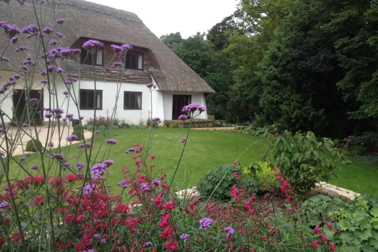 Backyard view of a thatched-roof home with lawn and vibrant flowering plants in the foreground, framed by trees.