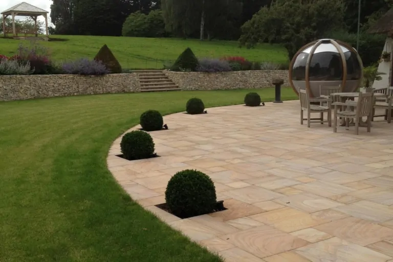 Paved patio with manicured box hedges and topiary balls, with a curved lawn and garden seating in the background.