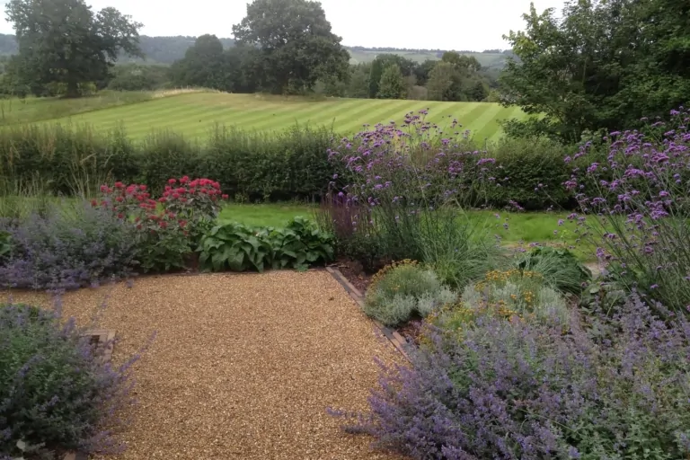 Gravel pathway lined with flowering plants leading to a scenic garden with a lawn and surrounding trees.