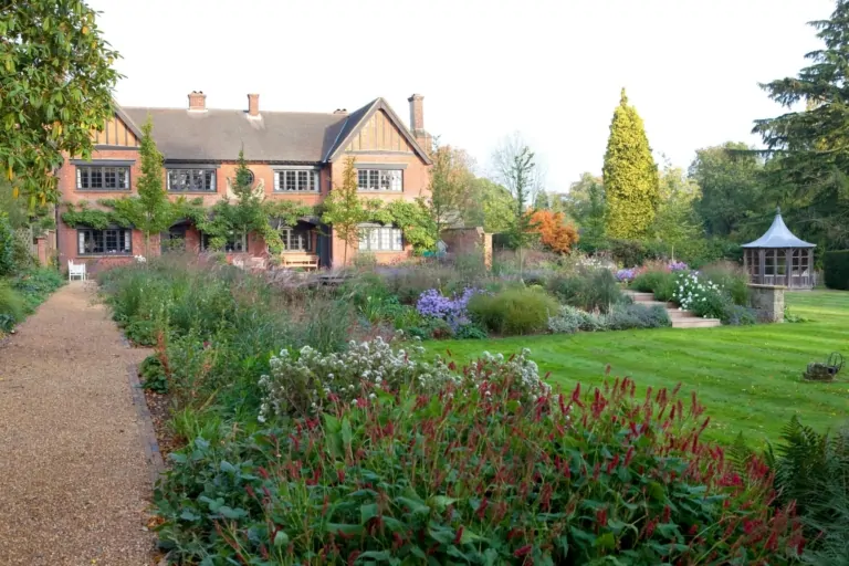 Elegant Surrey garden with lush perennial borders, manicured lawn, and traditional brick country house in the background, designed by We Love Plants.