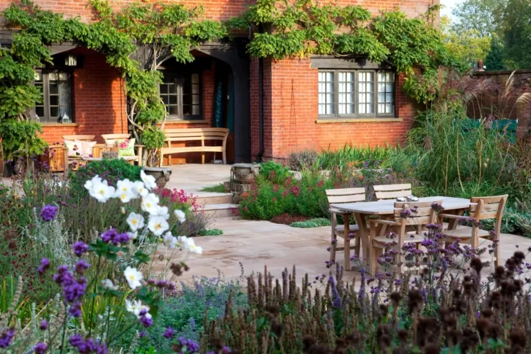 Close-up of paved garden terrace with wooden table and chairs, framed by mixed perennial plantings and vibrant blooms in a high-end Surrey garden.
