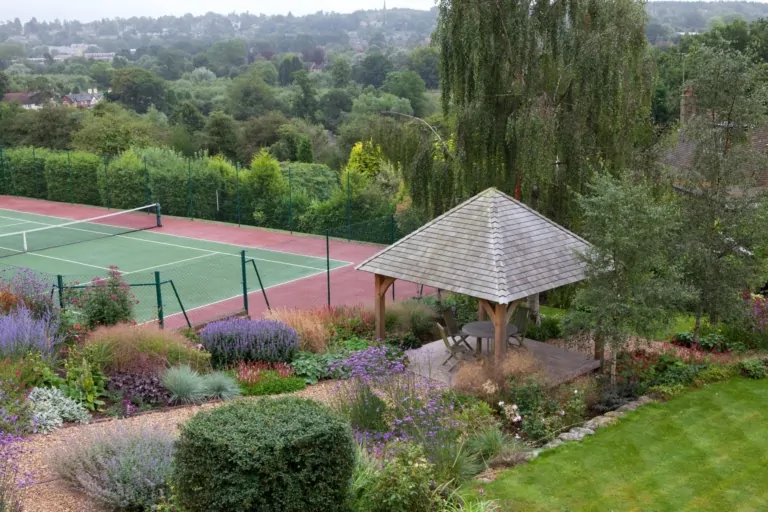 Tennis court in a landscaped garden with a wooden gazebo, surrounded by lavender, flowering plants, and neatly trimmed greenery.