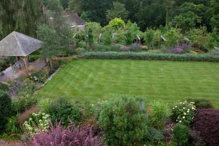 Manicured lawn with surrounding flowering borders and trees, viewed from an elevated perspective in a spacious garden.