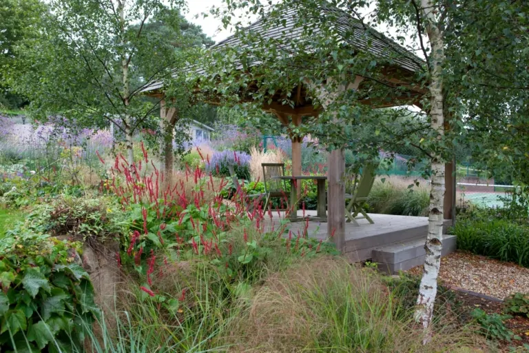 Wooden gazebo surrounded by lush garden plants, tall grasses, and colorful flowers creating a peaceful outdoor retreat.