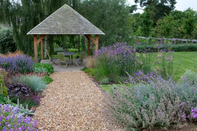 Gravel pathway leading to a wooden gazebo in a landscaped garden with lavender, flowering plants, and neatly trimmed shrubs.