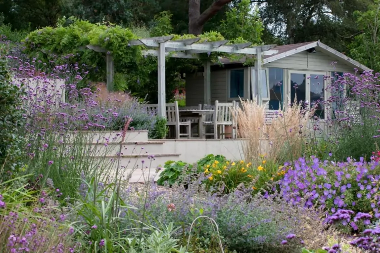 Outdoor dining area with white table and chairs set on a terrace, surrounded by lush flowering plants and garden greenery.