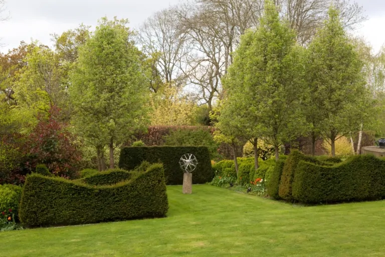 Formal garden with neatly trimmed hedges, green lawn, and sculptural centerpiece surrounded by trees in a landscaped estate.