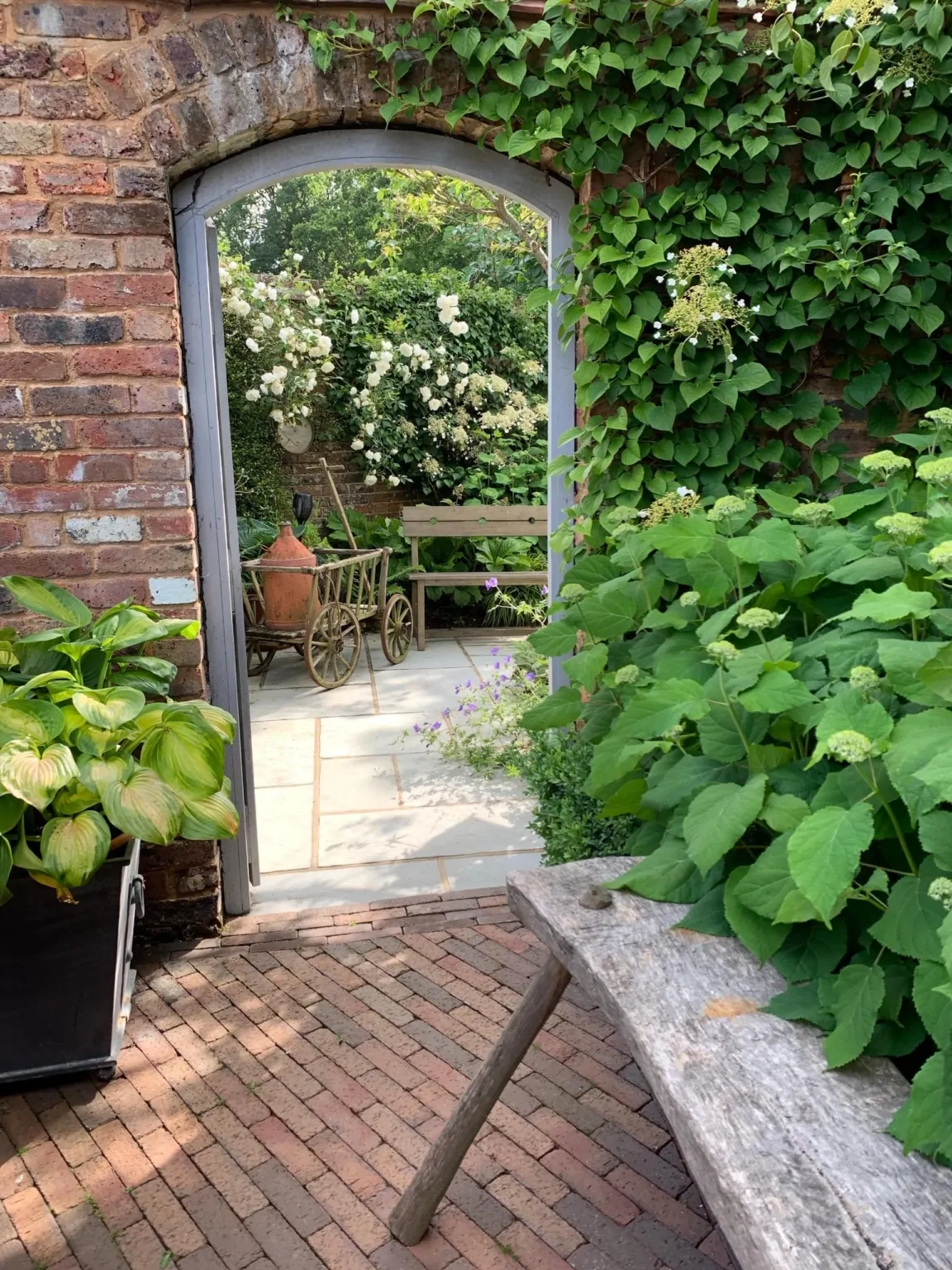 Stone garden terrace viewed through an arched brick doorway, surrounded by lush green foliage and potted plants, illustrating a tranquil and intimate Surrey garden design.