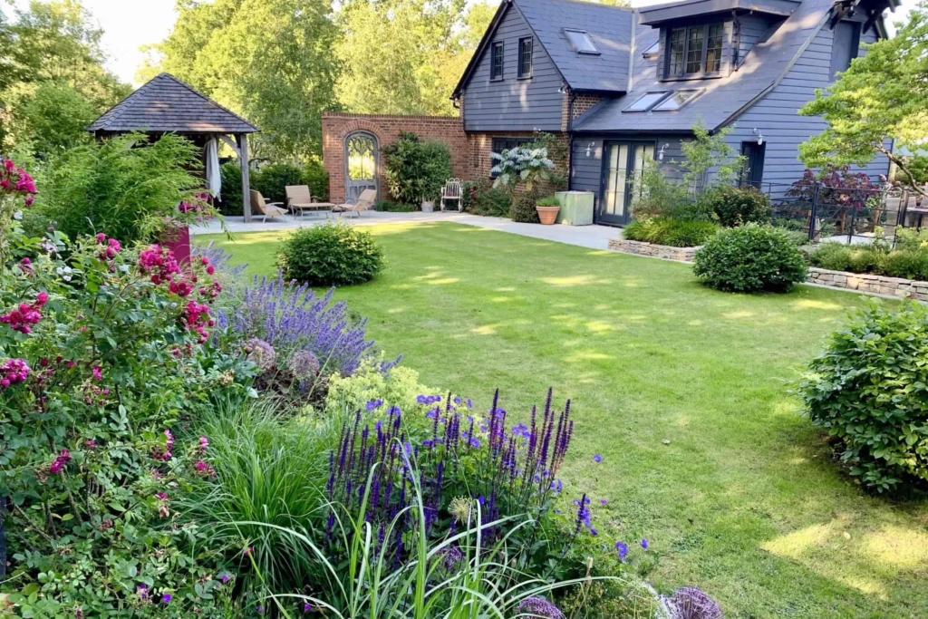 Contemporary house with dark slate exterior overlooking a landscaped garden with manicured lawn, flowering borders, and a wooden gazebo, creating a serene backyard retreat.