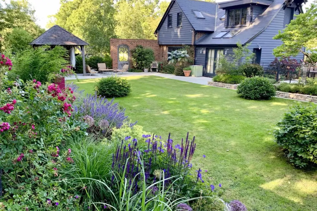 Contemporary house with dark slate exterior overlooking a landscaped garden with manicured lawn, flowering borders, and a wooden gazebo, creating a serene backyard retreat.