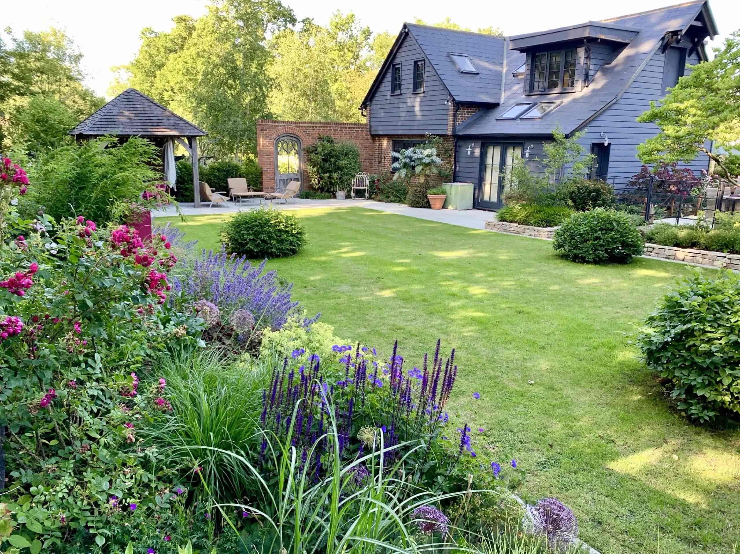 Contemporary house with dark slate exterior overlooking a landscaped garden with manicured lawn, flowering borders, and a wooden gazebo, creating a serene backyard retreat.