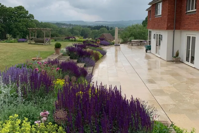 Surrey garden showing a stone terrace with layered perennial planting in purple, red, and green, leading the eye toward distant hills and a manicured lawn.
