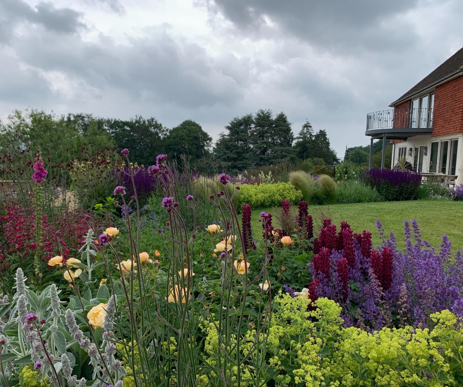 Wide Kent garden view with layered flower borders of red, purple, and yellow perennials, manicured lawn, and a distant brick building under a cloudy sky.