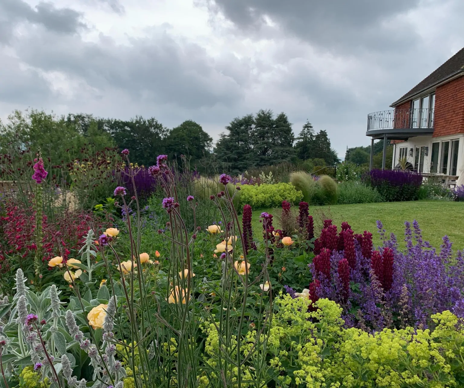 Wide Kent garden view with layered flower borders of red, purple, and yellow perennials, manicured lawn, and a distant brick building under a cloudy sky.
