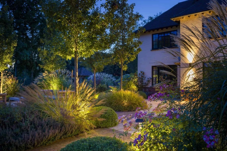 Twilight garden with pathway lighting, manicured shrubs, ornamental grasses, and contemporary home in the background.