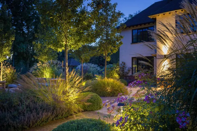 Twilight garden with pathway lighting, manicured shrubs, ornamental grasses, and contemporary home in the background.