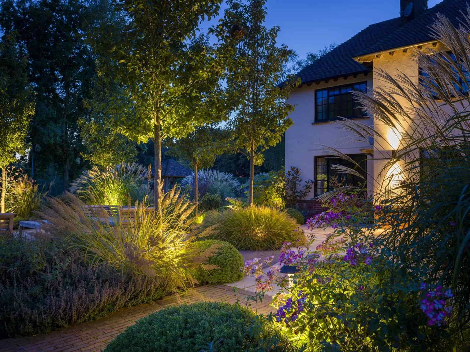 Twilight garden with pathway lighting, manicured shrubs, ornamental grasses, and contemporary home in the background.