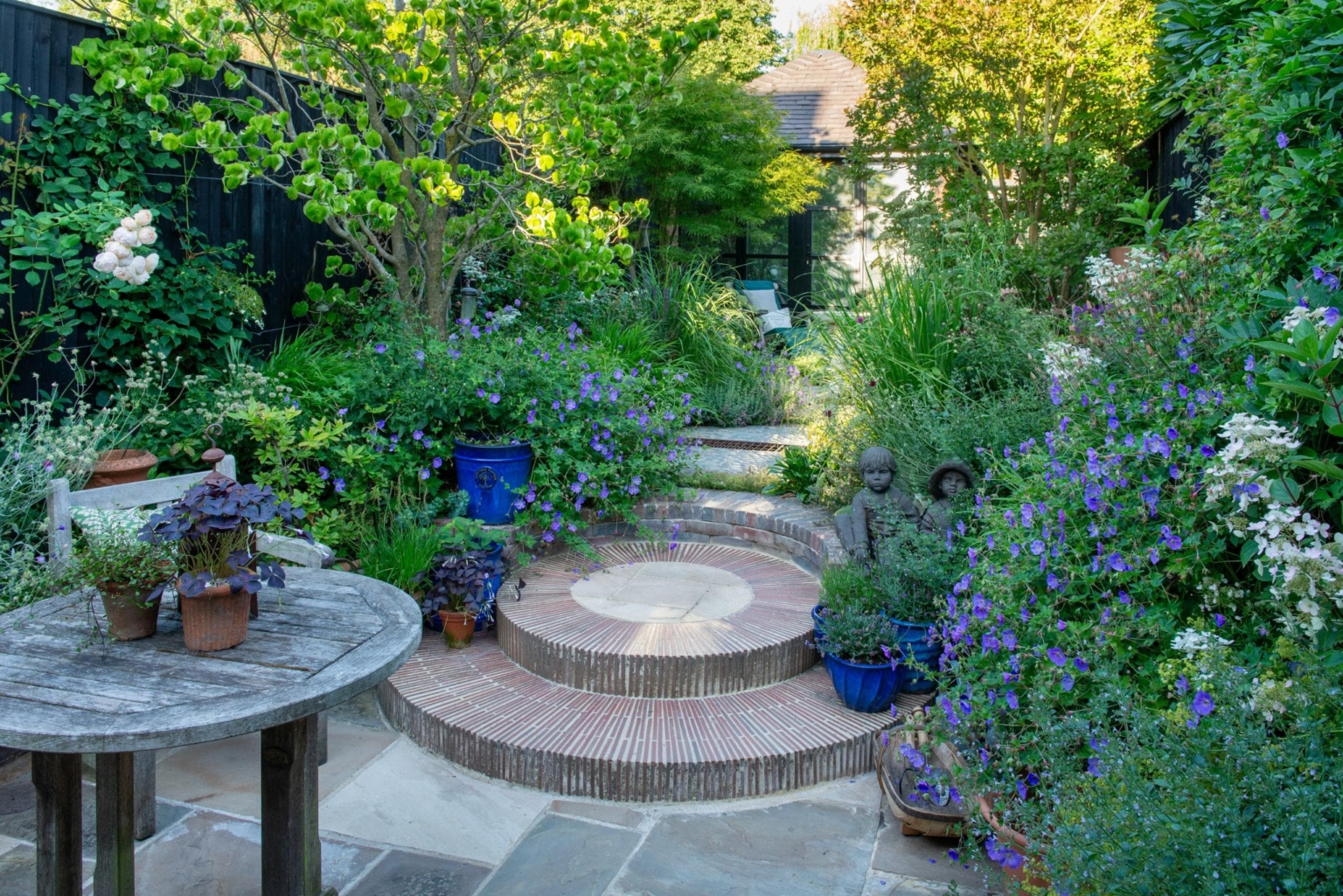 Circular paved garden terrace with seating, surrounded by vibrant perennial planting and shaded greenery, demonstrating high-end garden design.