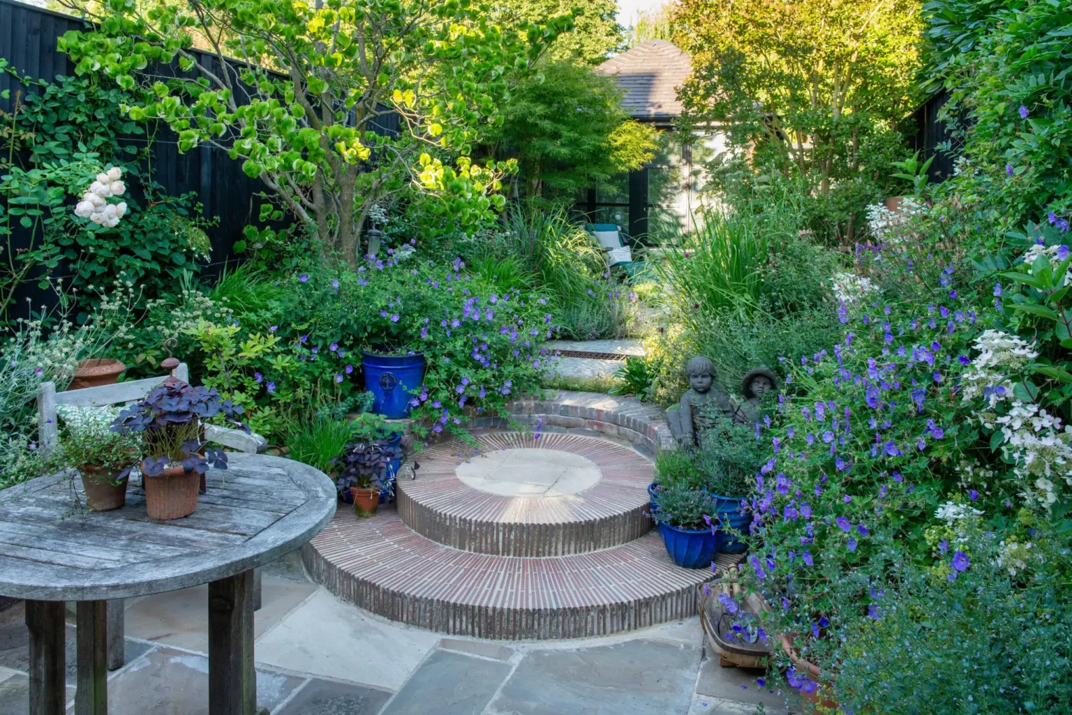 Circular paved garden terrace with seating, surrounded by vibrant perennial planting and shaded greenery, demonstrating high-end garden design.
