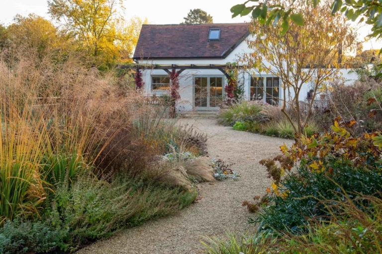 Autumn garden with gravel pathways, ornamental grasses, and a white house in the background creating a seasonal landscaped retreat.