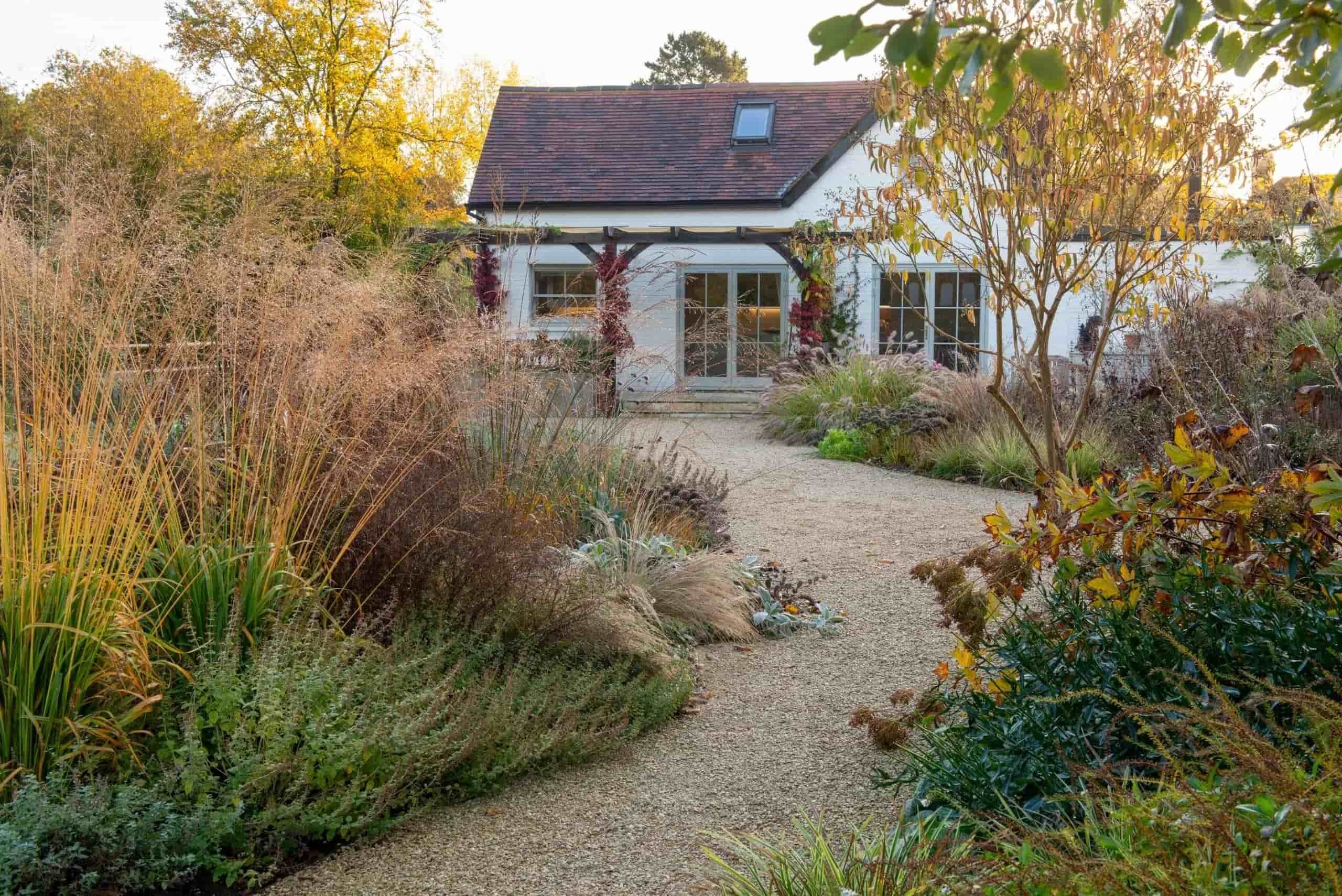 Autumn garden with gravel pathways, ornamental grasses, and a white house in the background creating a seasonal landscaped retreat.