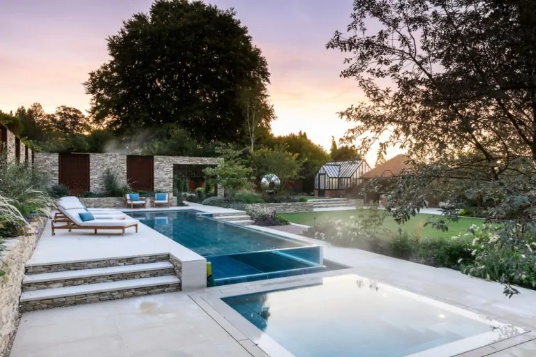 Luxury backyard with swimming pool, stone steps, landscaped garden beds, and contemporary seating under a tree-lined horizon.
