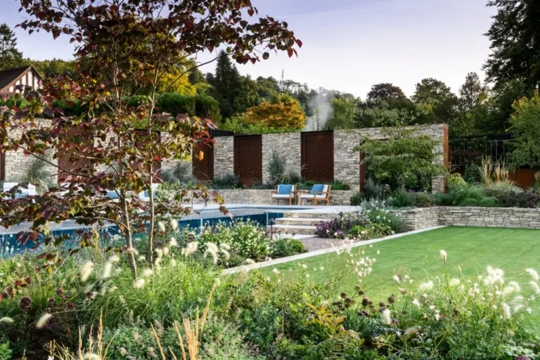 Luxury home with backyard pool, paved terrace, outdoor seating, and lush garden beds with ornamental plants.