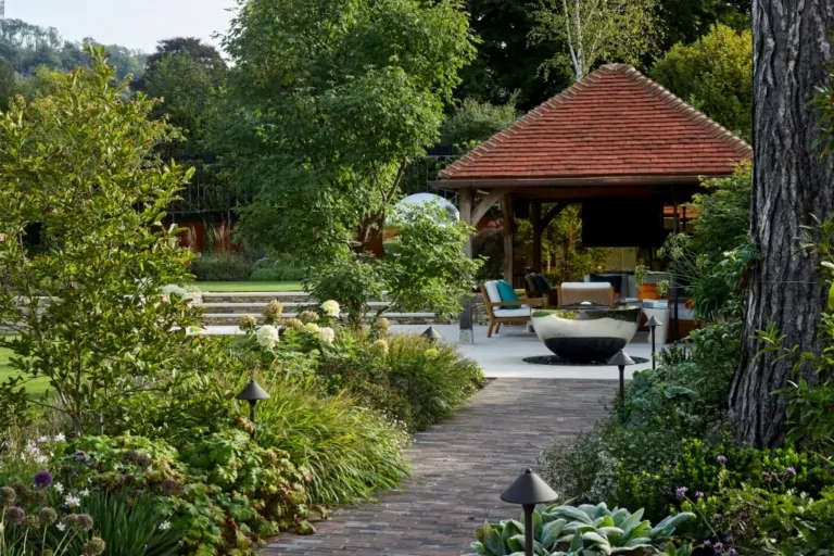 Garden path with ornamental shrubs, flowering borders, and a wooden gazebo leading to a poolside seating area.