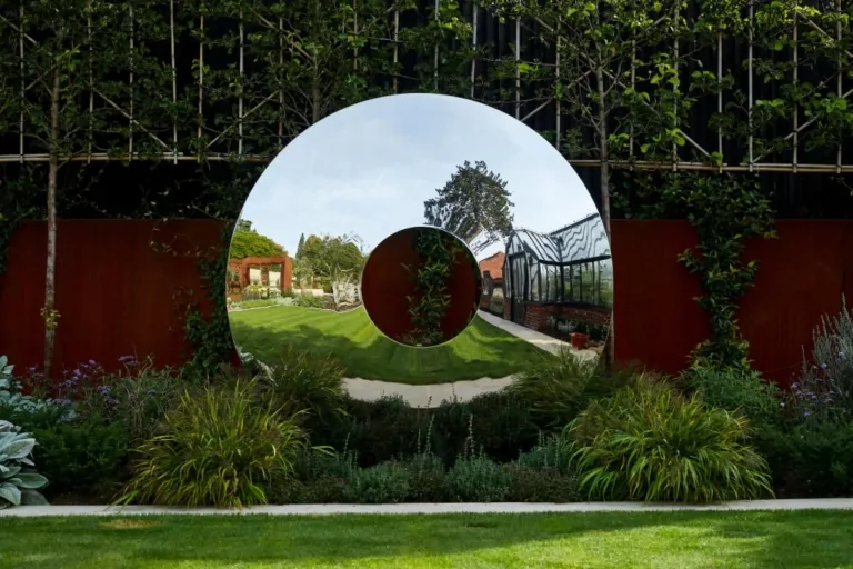 Circular mirror wall in a landscaped garden reflecting lawn, flowering plants, and greenhouse structures, adding visual depth.