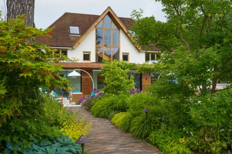Modern house with large windows and landscaped path bordered by dense green plants and ornamental shrubs.